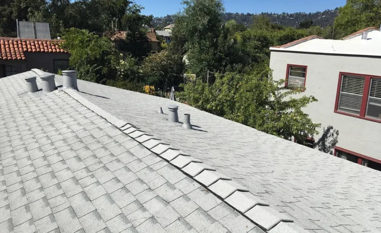 A gray shingle roof with three vents and ridge caps under a clear blue sky, overlooking distant houses and trees.